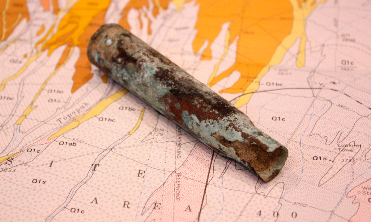 An empty shell casing from the bullet of a large gun, showing signs of various oxidations, sitting on top of a geologic map.