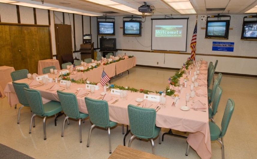 An institutional dining room with a table and chairs arranged in a U-shape around a projector.